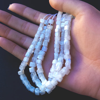 A person holds delicate strands of White Rainbow Moonstone beads, showcasing their smooth cube design.